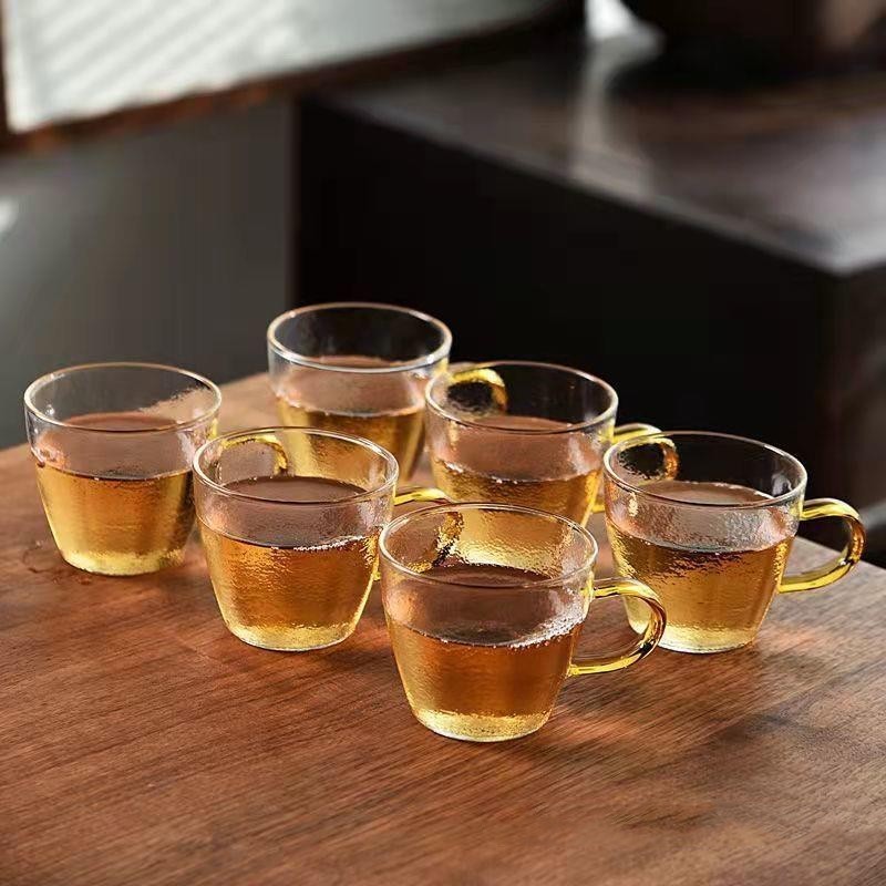 A beautifully arranged Thick Glass Transparent Tea Set, featuring a minimalist design with high borosilicate glass. The set includes six elegant tea cups and a matching cup holder, perfect for serving tea. Available in various sizes and styles, including transparent and colorful options with long spout pots.