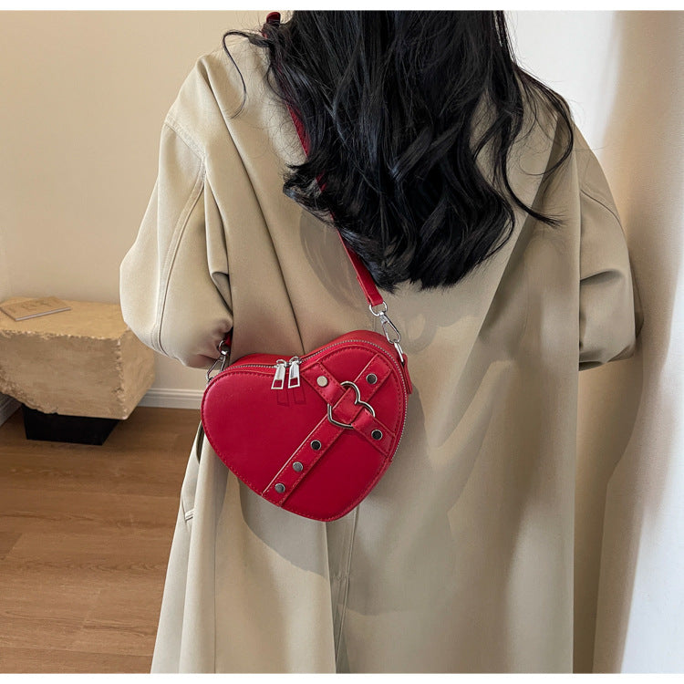 Cute heart-shaped shoulder messenger bag made of soft PU material, featuring a zipper change bag inside. Available in black, cream white, silver, and wine red, with a single shoulder strap and an urban simplicity style for everyday use.